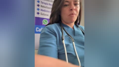 A video of a female nurse with long brown hair, wearing a light blue scrub top, stethoscope around her neck, in a hospital setting with a blurred background.