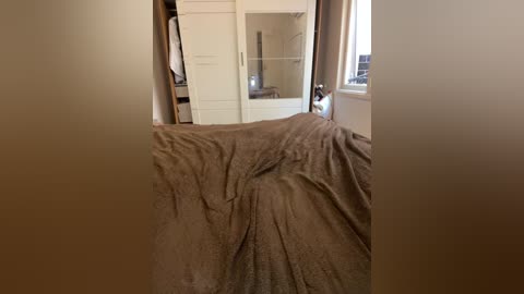 Video of a bedroom with a messy, unmade bed covered in a beige comforter, white built-in wardrobes, and a window with beige curtains, creating a cozy, uncluttered atmosphere.
