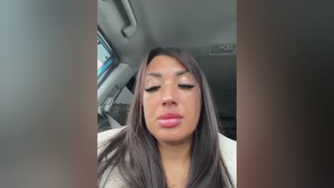 Video of a young woman with long, straight dark hair, wearing makeup and a white top, seated in a car with a gray interior.