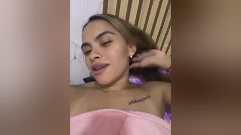 Video of a young woman with light skin, long brown hair, and closed eyes, lying on her back, wearing a pink strapless top, with a tattoo on her collarbone.