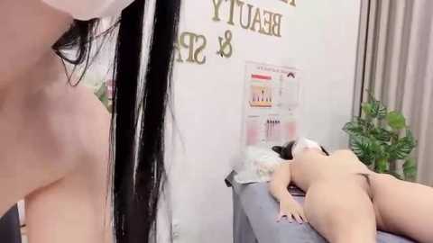 Video of a nude woman with fair skin lying face down on a massage table in a dimly lit room with posters and a plant in the background.