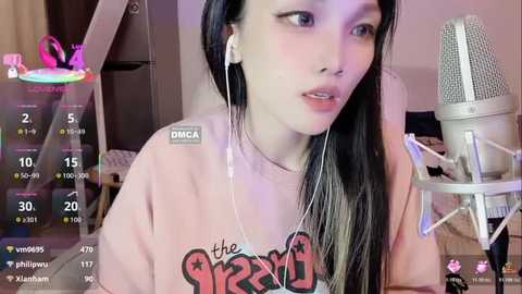 Video of an Asian woman with long black hair, wearing a pink \"the king\" t-shirt, white headphones, and a microphone. Background shows a pink butterfly decoration and a grey chair.