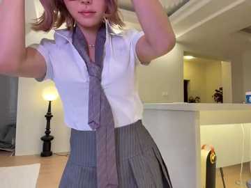 Video of a woman in a white blouse and gray pinstriped skirt, holding a purple tie, in a modern, well-lit living room with white walls and wooden floors.