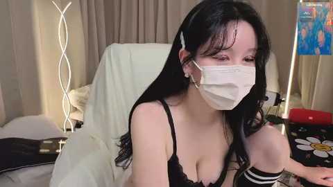 A video of a young Asian woman with long black hair and a white face mask, wearing a black lace bra and black thigh-high socks. She's in a dimly lit room with a bed and a colorful poster in the background.