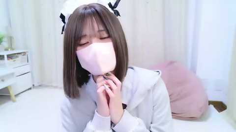 Video of a young East Asian woman with straight, shoulder-length brown hair, wearing a white sailor hat, pink face mask, and a white jacket, kneeling on a white floor, holding hands, in a minimalist, bright room.