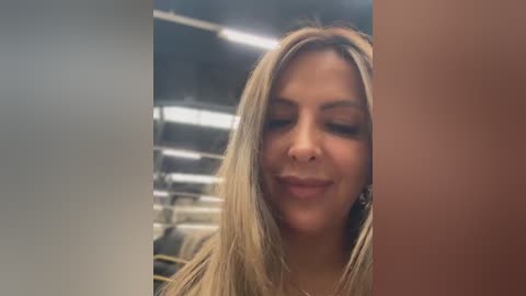 A video of a smiling woman with long blonde hair, wearing a black top, captured indoors with fluorescent lights and a blurred staircase in the background.