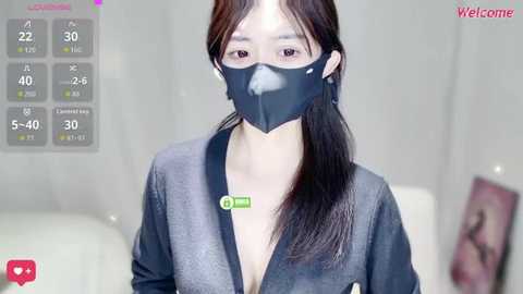 Video of a young East Asian woman with long black hair, wearing a dark mask, gray robe, and green badge, in a modern, bright room with a white sofa.