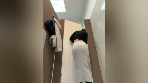 Video of a woman with a curvy figure in a tight black top and white skirt, washing her hands in a narrow bathroom with beige walls and a fluorescent light above.