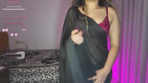 Video of a woman in a black sari, red bra, and long hair, standing in a dimly lit room with purple lighting, white walls, and a bed with a black and white patterned sheet.