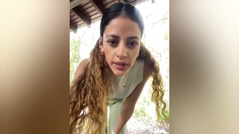 A video of a young woman with long, wavy, light brown hair styled in pigtails, wearing a light-colored sleeveless top, leaning forward with a serious expression. The background features a wooden structure with greenery outside.