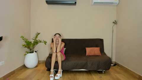 A video of a young woman with long dark hair, wearing a pink dress and white heels, sitting on a dark brown couch, hugging her knees, in a minimalist room with light wood floors and beige walls.