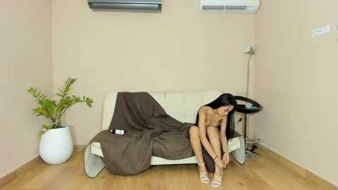 Video of a nude woman with long black hair tying a shoe on a beige couch in a minimalist room with wooden floor, brown blanket, white pot, and air conditioner.