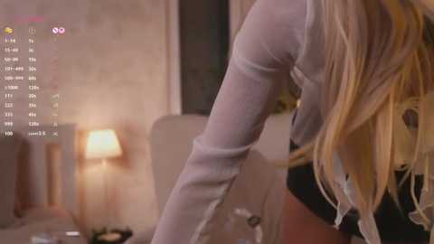 A video of a woman with long, blonde hair, wearing a sheer white top, bending over a table in a dimly-lit, cozy room with a lamp and abstract art on the wall.