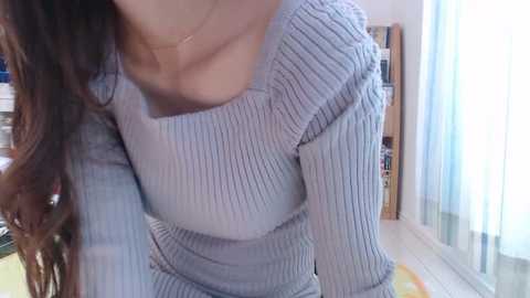 Video of a woman with long, wavy brown hair wearing a ribbed, light blue, off-the-shoulder sweater, indoors near a window with sheer curtains.