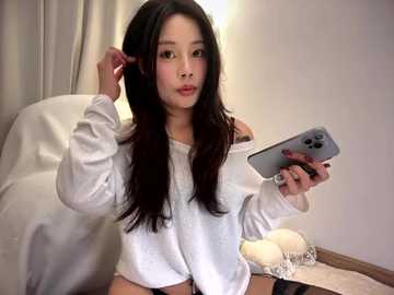 Video of an East Asian woman with long black hair, fair skin, and light makeup, wearing a white off-shoulder sweater, holding a smartphone. Background includes a white couch, beige curtain, and wooden floor.