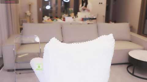 A video of a modern living room with a white, textured throw pillow, beige sofa, and round glass table. The background features a cluttered dining area with a glass door and a mirror.