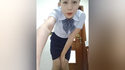 A video of an East Asian woman with fair skin, dressed in a white blouse and navy skirt, bending over, revealing her underwear. She stands in front of a wooden door.