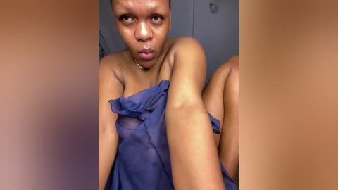 Video of a dark-skinned woman with short hair, wearing a sheer blue dress, sitting in a dimly lit room with blurred background.