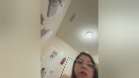 A blurry, low-resolution video of a woman with long brown hair, wearing glasses, standing in a beige-tiled bathroom with a white mirror and a round light fixture.