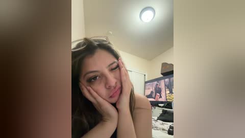 A video captures a young woman with long dark hair, resting her face in her hands, winking, in a dimly lit room with a TV displaying a movie scene.