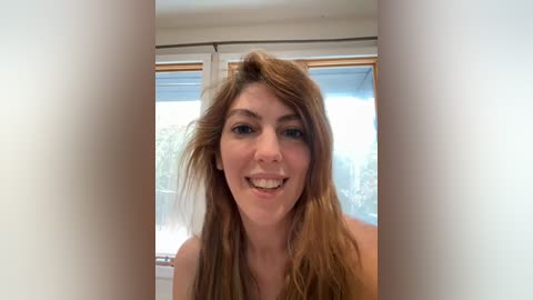 A video of a young Caucasian woman with long, light brown hair, fair skin, and a friendly smile. She is indoors near a window with a white curtain.