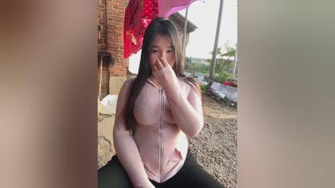 Video of an Asian woman with long black hair, wearing a pink zip-up hoodie, hiding her face with her hand, standing outside near a brick building with a red umbrella.