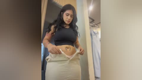 Video of a Latina woman with long black hair, tattoos, wearing a black crop top and beige pants, adjusting her pants in a dimly lit hallway.