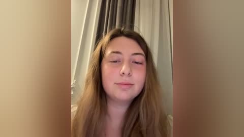Video of a young woman with long, light brown hair, fair skin, and closed eyes, standing in a dimly lit room with gray curtains and beige walls.