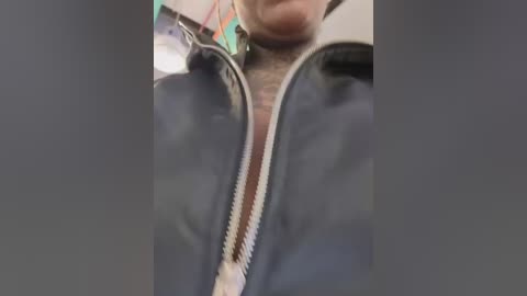 A close-up video of a person wearing a black leather jacket with a zipper down the center, revealing a light-colored shirt underneath. The background is blurred, featuring indistinct objects and colors.