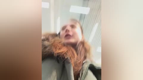 A blurred video shows a young woman with light skin, long reddish-brown hair, and a green jacket with a fur collar, standing in a brightly lit room with vertical blinds.