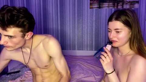 A video of a shirtless young man with a slender physique and a slender young woman with straight brown hair, both nude, in a dimly lit bedroom with a purple and blue striped wall and a framed picture.