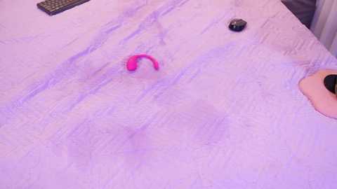 A video showing a light purple bedspread with a pink, ring-shaped silicone sex toy and a black remote control on the left, and a man's hand with a black watch on the right.