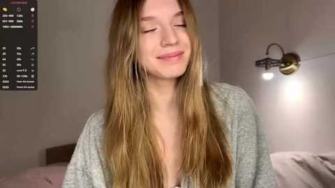 Video of a young Caucasian woman with long, straight blonde hair, wearing a light grey cardigan, smiling softly, in a dimly lit bedroom with a bed and wall-mounted light.