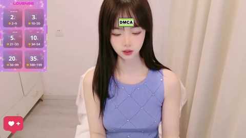 Video of an Asian woman with long black hair, fair skin, wearing a light blue, quilted dress, sitting in a minimalistic room with white walls and a cream curtain.