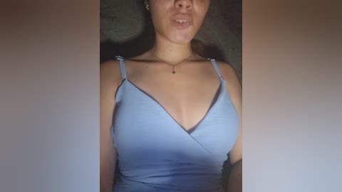 A video of a light-skinned woman with medium-length brown hair, wearing a light blue V-neck camisole. She has a slender build and a small, delicate necklace. The background is a dark, textured surface.