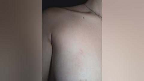 Video of a fair-skinned person's upper torso, showing a small mole on the right shoulder and a thin silver necklace. The background is a plain, beige wall.