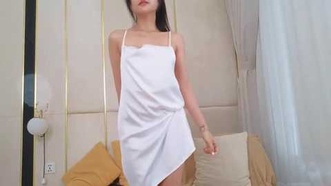 Video of an Asian woman with long black hair, wearing a white satin slip dress, standing in a modern bedroom with beige walls, a gold lamp, and a bed with yellow pillows.