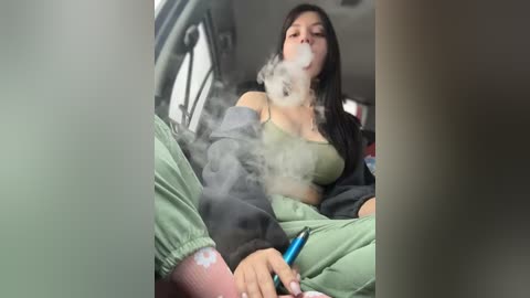 A video of a woman with long black hair, wearing a green top and green pants, sitting in a car, smoking a cigarette with visible smoke.