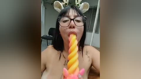 A video shows a topless Asian woman with glasses, wearing a floral headband, licking a large, spiral-shaped lollipop with orange and pink stripes. The background features a gray room with a chair and a door.