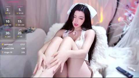 A video of an East Asian woman with long black hair, wearing white lingerie and a white headband, sitting on a plush white couch in a softly lit room.