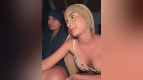 Video of two women seated close together, one with a turban and the other in a white lace bra, both with fair skin and light makeup, looking forward.