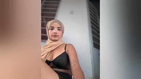 Video of a light-skinned woman with a light beige hijab and black bra, sitting on a staircase.