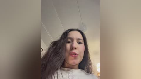 A video of a woman with long, dark hair and fair skin, wearing a white top, against a beige wall with visible ceiling beams. She has a neutral expression and is slightly out of focus.