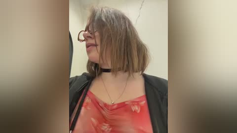 Video of a fair-skinned woman with shoulder-length brown hair, wearing glasses, a black choker, and a red floral blouse. She stands against a plain, off-white background.