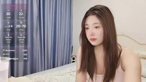 Video of a young Asian woman with long brown hair, wearing a pink tank top, sitting on a bed with light-colored bedding. The background shows a blue curtain and a digital screen displaying weather information.
