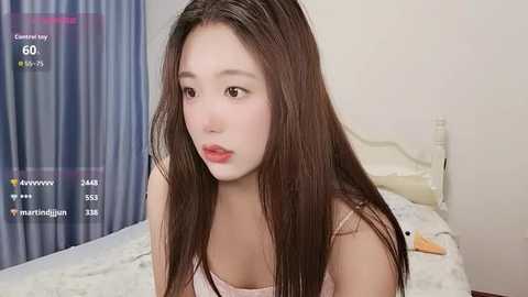 Video of a young Asian woman with long brown hair, fair skin, and red lipstick, wearing a white tank top, sitting on a bed with blue curtains in the background.