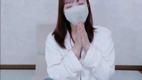 A video of a young woman with light skin, wearing a white surgical mask, white robe, and a blue top, hands clasped in prayer, seated in a plain white room.