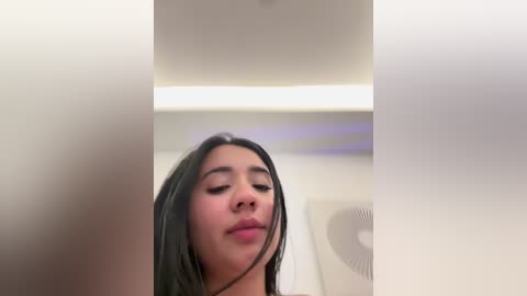 A video of a young woman with long, dark hair, closed eyes, and a neutral expression. The background features a minimalist room with a ceiling fan and a strip of light.