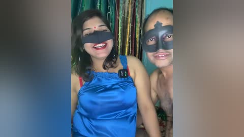 Video of two smiling women with black masks over their eyes, wearing blue satin tops, standing in front of a colorful, vertical cloth backdrop.