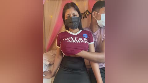 Video of a woman with a black face mask, wearing a maroon \"JATE AIRWAYS\" T-shirt and black skirt, being held by a man in a pink shirt, in a dimly lit room with pink curtains.